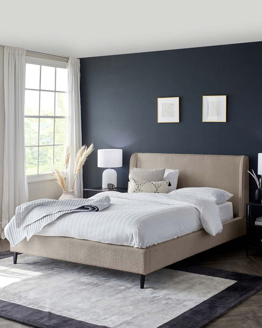 Elegant contemporary bedroom showcasing a large upholstered bed with a high, tufted headboard in a neutral beige fabric, accompanied by crisp white bedding and accent pillows with various textures. A sleek bedside table with a modern white lamp complements the bed. The room is grounded by a plush, two-tone grey area rug.