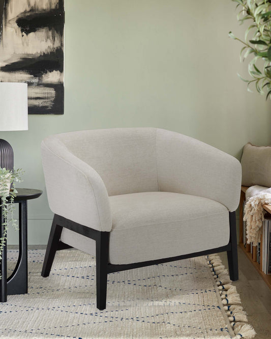 Modern barrel chair with a curved, textured light grey upholstery and contrasting black wooden legs, displayed in a stylish interior with a decorative textured rug, adjacent to a sleek black round side table with a potted plant atop, and near a bookshelf with assorted items. A large abstract black and white painting hangs on the wall, while a white lamp with a fabric shade stands in the background.