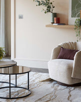 Modern living room featuring a round, black-framed glass-top coffee table and a cosy beige upholstered armchair with a textured throw pillow. An ornamental floating wooden shelf is mounted above, adorned with potted greenery and decorative objects.