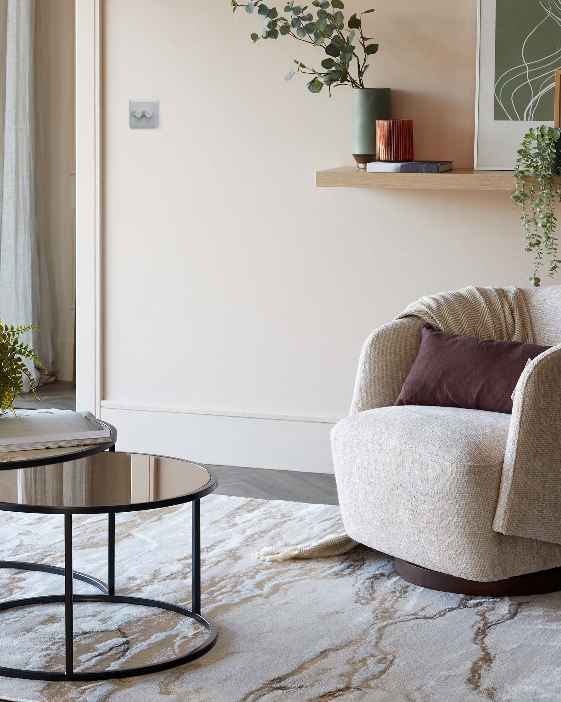 A cosy, contemporary beige armchair with textured upholstery and plush cushions paired with a modern round side table featuring a sleek, black frame and reflective tabletop. A light wooden shelf is mounted on the wall above, adorned with decorative items. The furniture rests on an elegant, cream-colored area rug with subtle marbling.