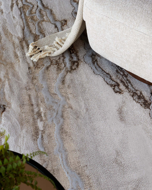 Part of a neutral-toned fabric sofa is visible at the upper right portion of the image, showing a glimpse of its textured upholstery and a cushion. Below the sofa, an area rug with intricate wavy patterns in shades of cream, beige, and light brown extends across the floor, partially obscured by a cream-colored woven throw with fringe detail that hangs over the edge of the sofa.