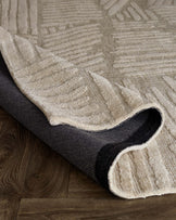 Beige textured area rug with a contemporary leaf pattern, partially rolled to show the black underside, on a wooden floor.