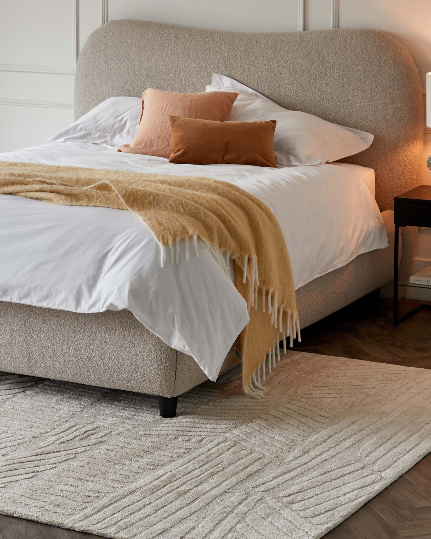 Contemporary upholstered bed with a curved headboard in beige fabric, styled with white bed linen, cinnamon-coloured accent pillows, and a caramel throw blanket. Beside the bed is a small dark wood nightstand with an open shelf, resting on a textured light-coloured area rug.