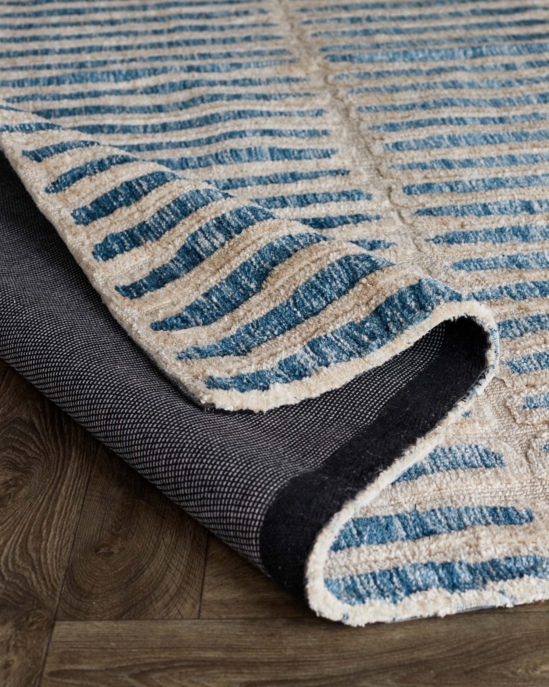 Textured area rug with beige base and blue stripe pattern, partially rolled up to reveal a black anti-slip underlay, on a wooden floor.