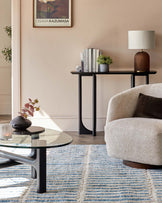 Contemporary living room showcasing a round glass coffee table with a unique black base, a sleek black console table adorned with books and a modern lamp, and a cosy beige armchair with a dark throw pillow. A striped blue and white area rug anchors the space.