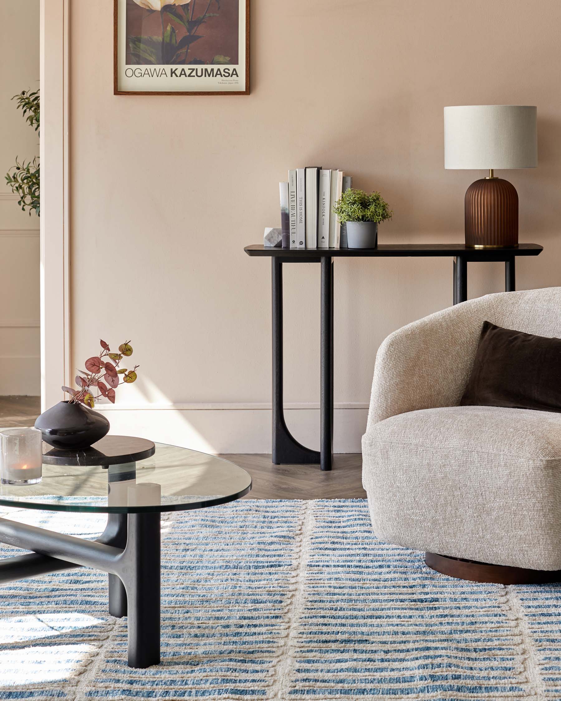 Contemporary living room showcasing a round glass coffee table with a unique black base, a sleek black console table adorned with books and a modern lamp, and a cosy beige armchair with a dark throw pillow. A striped blue and white area rug anchors the space.