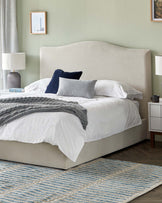 An elegant upholstered bed with a curved headboard and neutral-toned fabric, featuring white bedding with a variety of pillows including grey and navy blue accents, complemented by a soft grey knit throw. Beside the bed is a sleek white nightstand with a grey lamp and a silver base, and underneath is a textured blue and beige area rug.