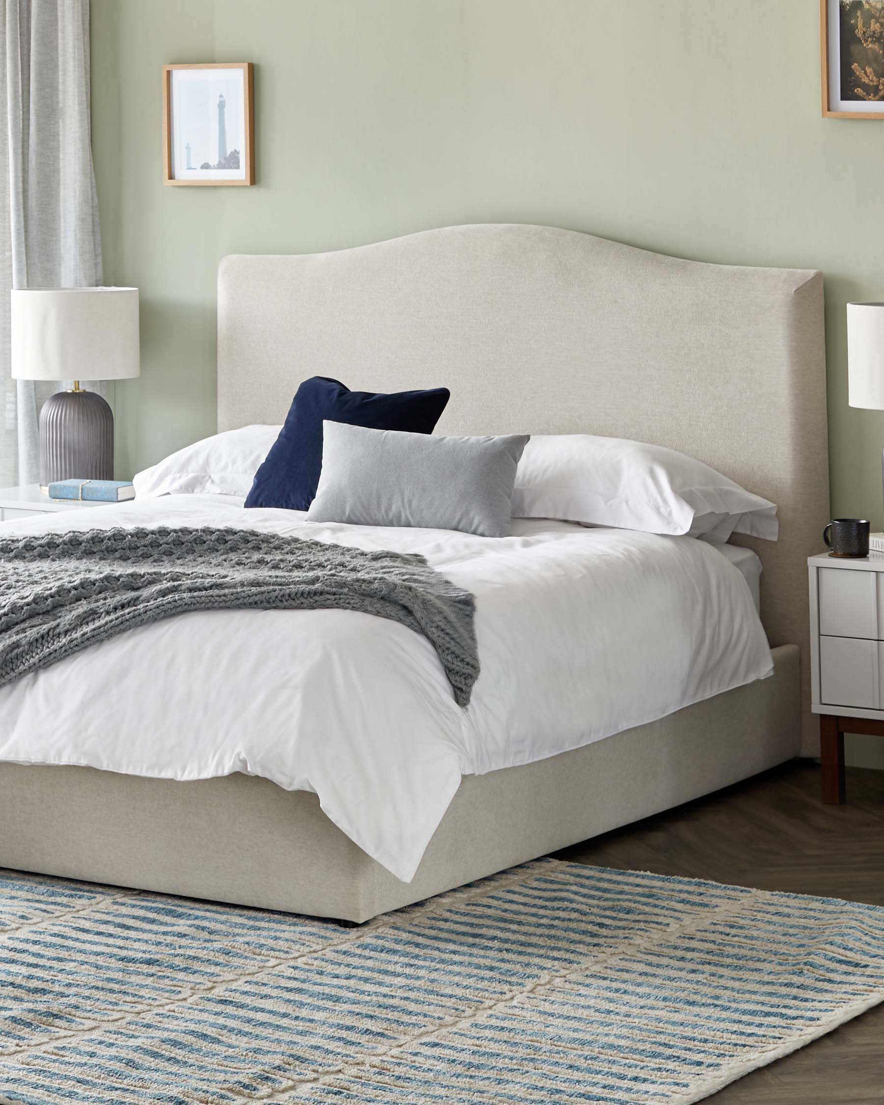 Elegant bedroom furniture set featuring an upholstered king-sized bed in a neutral beige fabric with curved corners on the headboard, flanked by two contemporary white nightstands with sleek metallic handles. A textured blue and beige area rug rests on the floor, complementing the serene and sophisticated decor.