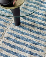 A contemporary round glass-top side table with a single black cylindrical leg atop a textured cream-colored area rug with a simple blue striped pattern. The table seems to have a thin gold rim adding a touch of elegance.