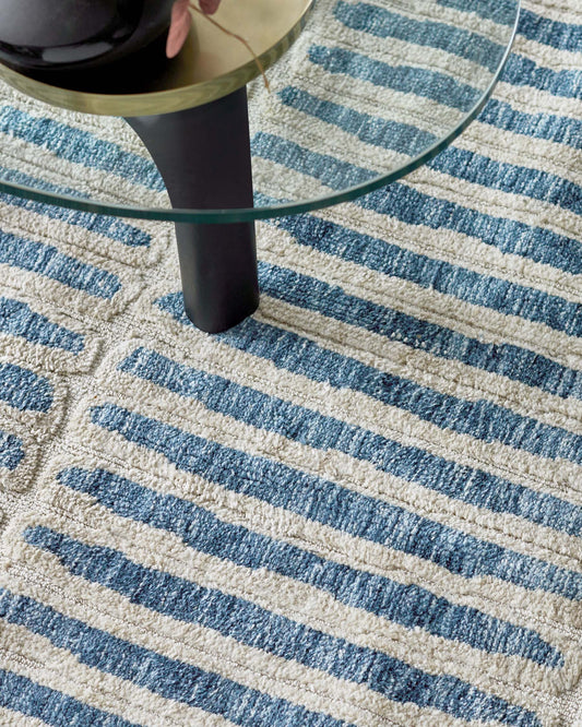 A contemporary round glass-top side table with a single black cylindrical leg atop a textured cream-colored area rug with a simple blue striped pattern. The table seems to have a thin gold rim adding a touch of elegance.