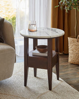 A stylish side table with a round marble top, dark wooden legs, and a shelf, featuring a glass of water and decorative bowl.