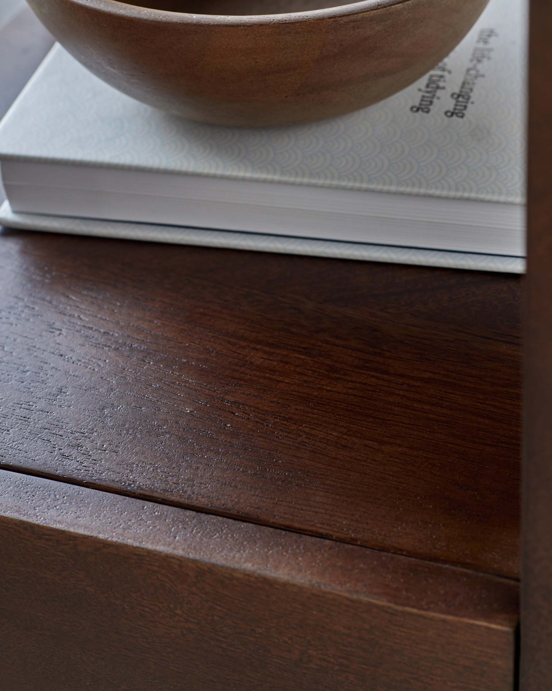 Dark wooden furniture surface with a subtle grain, topped with a decorative bowl and a patterned book.