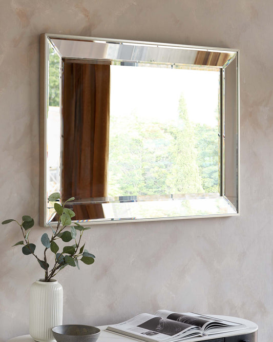 A contemporary room featuring a large square mirror with a wide, sleek metallic frame mounted on a textured wall. Below the mirror sits a cylindrical white ceramic vase with green foliage on a small, round dark table displaying an open magazine. There is also a small, shallow dark bowl beside the vase. The reflection in the mirror shows a scenic view of greenery outside the window.