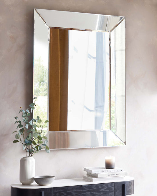 Modern black console table with a sleek and minimalistic design, featuring a smooth black surface with contrasting wood grain patterned drawers and discreet round knobs. The table is accessorized with a textured grey ceramic vase containing eucalyptus branches, two grey decorative bowls, a stack of hardcover books, and a lit scented candle. A large rectangular mirror with a simple, reflective frame hangs above the table, enhancing the bright and airy feel of the space.