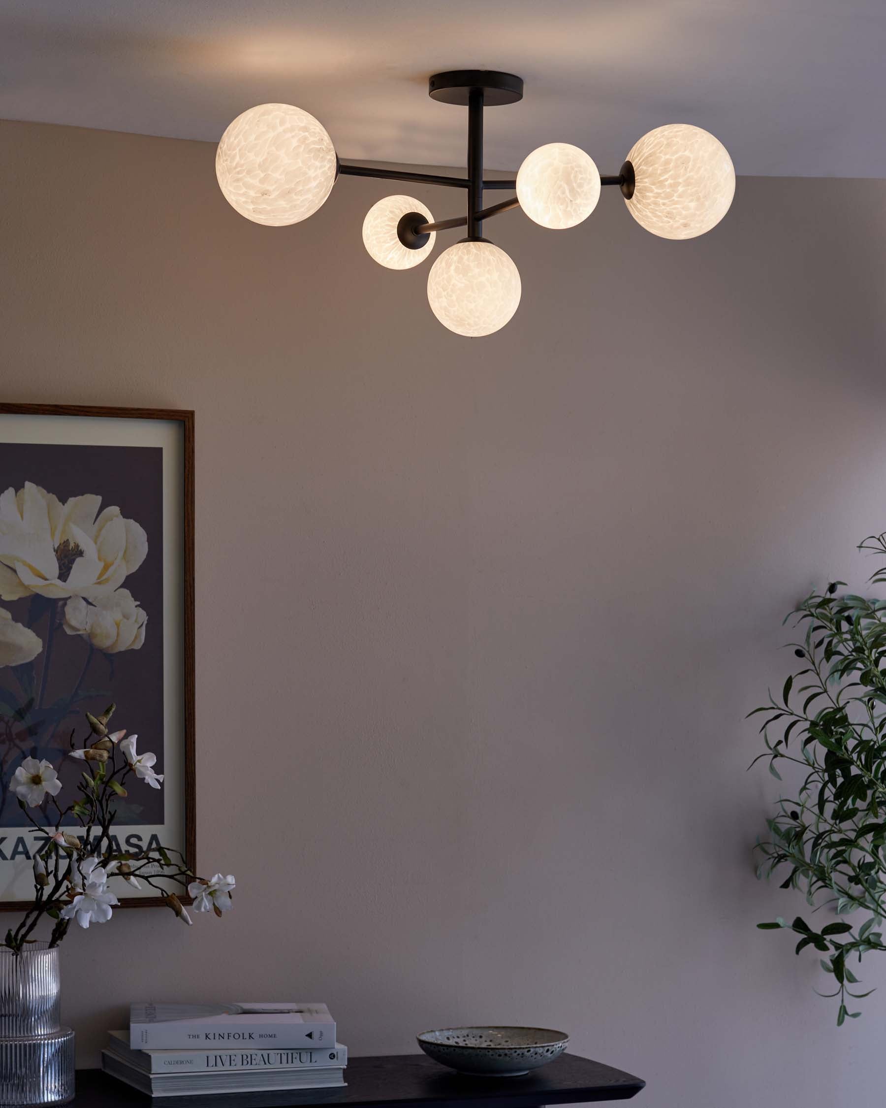 Modern minimalist black console table adorned with decorative items including a stack of hardcover books, a ceramic bowl, and a vase with white flowers. An artificial potted plant adds greenery to the scene. The setup is complemented by a contemporary chandelier with multiple frosted glass globes above.