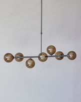 Modern linear pendant light with a series of spherical, smoked glass shades arranged along a horizontal metal bar, suspended from a ceiling mount by a sleek, adjustable cable.