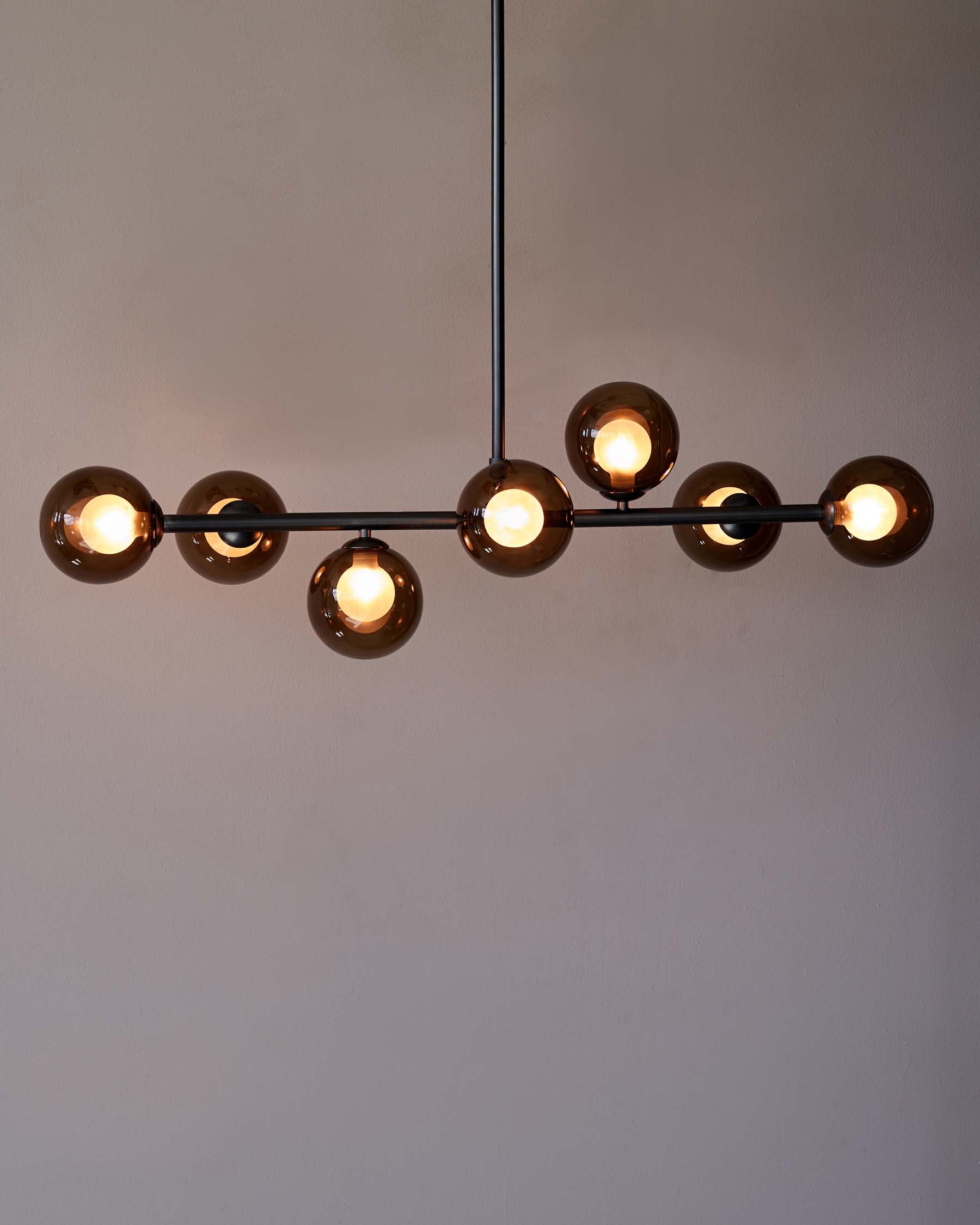 Modern linear pendant light with a black horizontal bar supporting six globular smoked glass shades, each encasing a lit bulb, suspended against a neutral background.