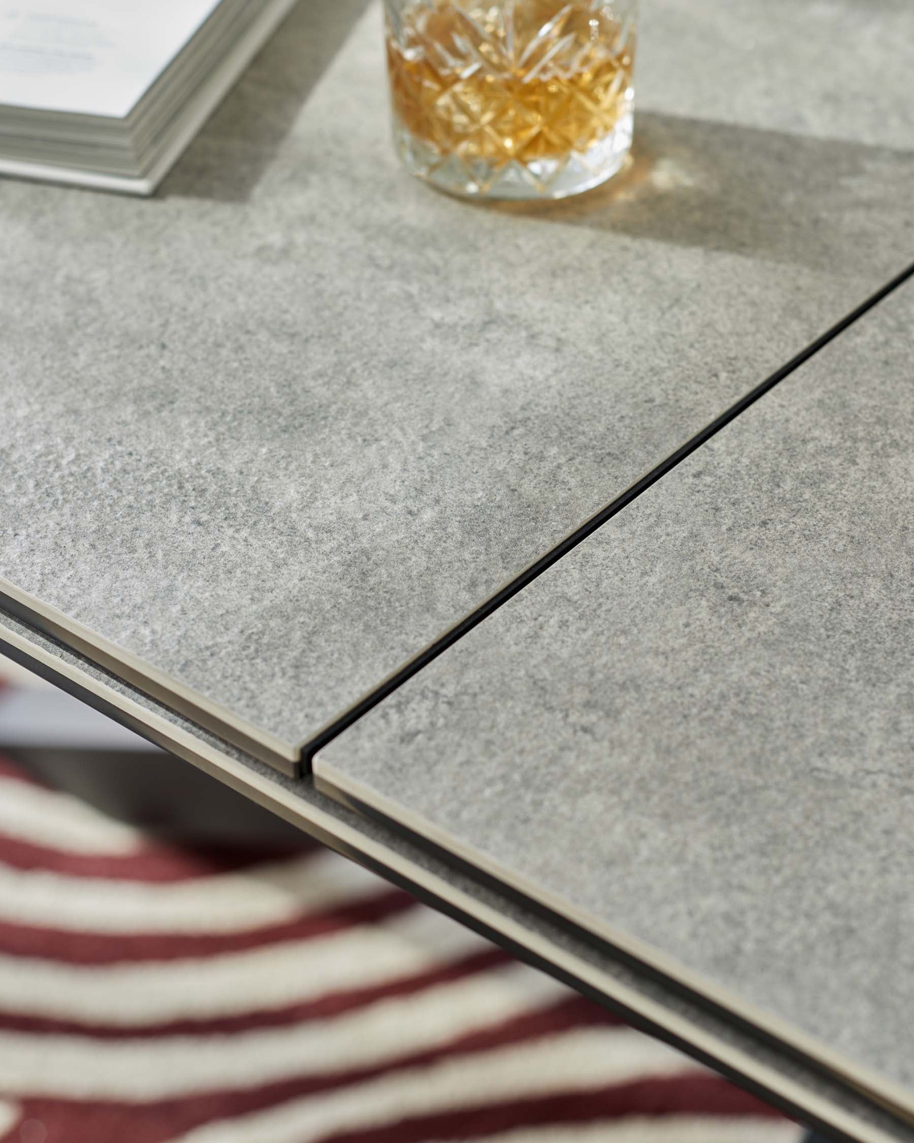 A close-up of a modern grey stone-textured table surface with sleek black lines separating the stone slabs and a minimalist metallic edge detail. A crystal glass partially filled with a golden liquid sits atop, beside an open book with visible text, all resting on a striped red and white fabric surface.