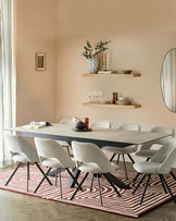 Modern dining room furniture set, consisting of a rectangular table with a concrete-style surface and black angular legs, complemented by six light grey upholstered chairs with black metal legs, arranged on a red and white patterned area rug. Two wooden floating shelves with decorative items are mounted on the wall above.