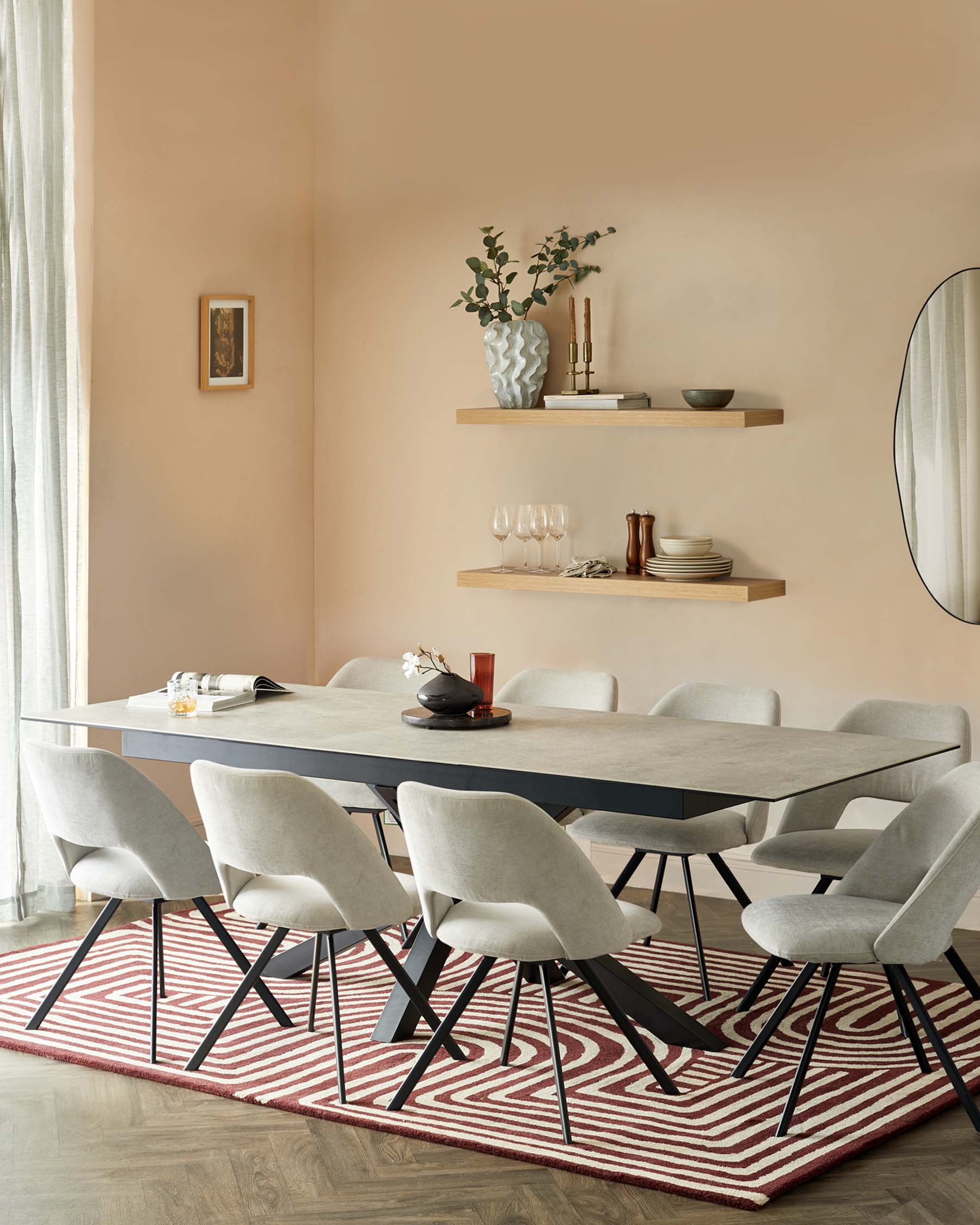 Modern dining room furniture set, consisting of a rectangular table with a concrete-style surface and black angular legs, complemented by six light grey upholstered chairs with black metal legs, arranged on a red and white patterned area rug. Two wooden floating shelves with decorative items are mounted on the wall above.