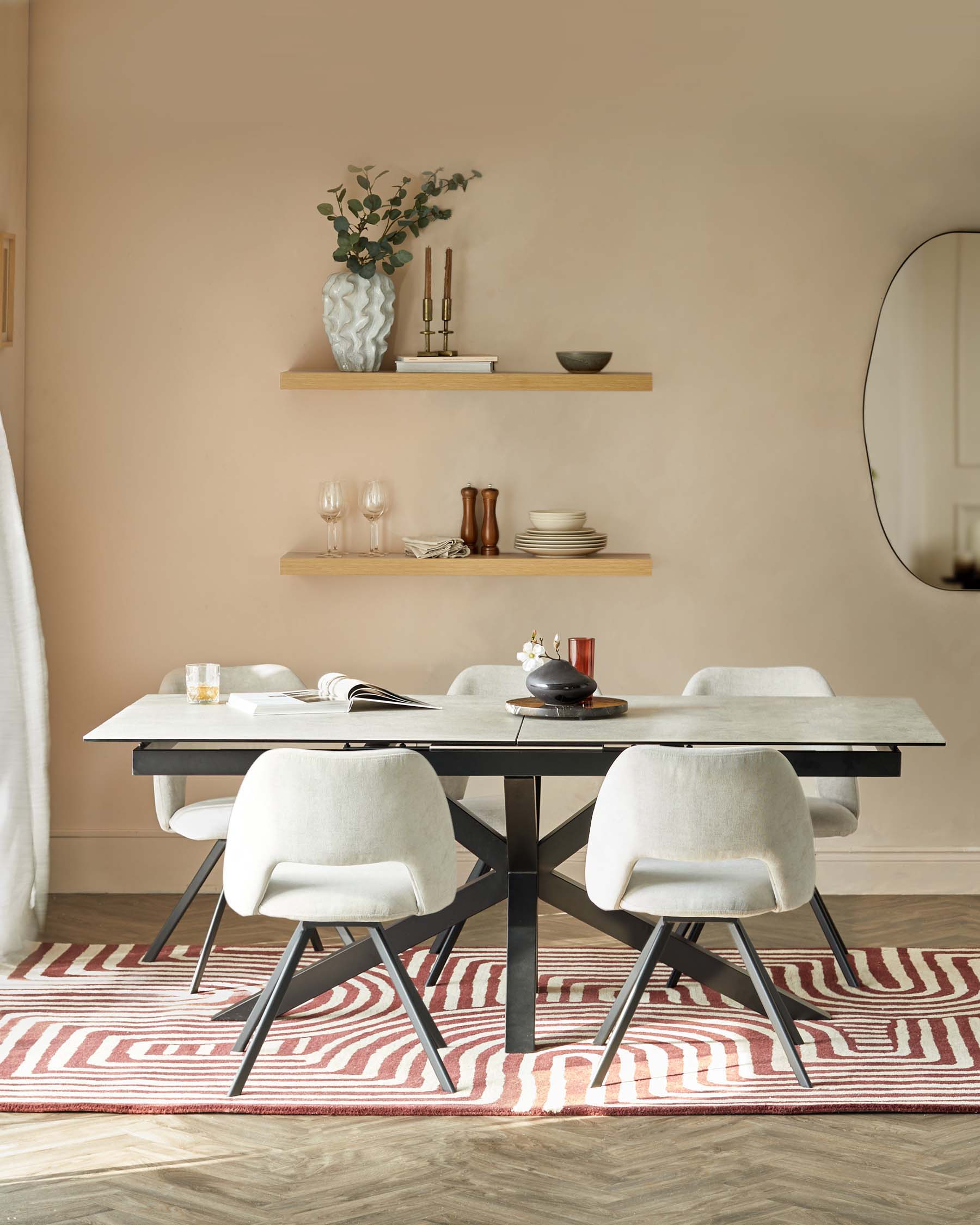 Modern dining room featuring a rectangular black table with a smooth surface on top of a striped red and beige area rug. Around the table, there are four light grey upholstered dining chairs with dark metal legs. On the wall behind the dining set are two wooden floating shelves displaying a selection of decorative items and tableware, and a large round mirror with a black frame is visible to the side, reflecting part of the room.