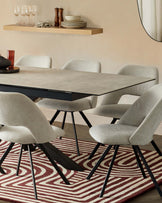 Modern dining room featuring a rectangular table with a textured light grey top and black metal legs arranged over a geometric red and white patterned area rug. Surrounding the table are six mid-century inspired chairs with plush light grey upholstery and black metal legs that match the table. The setting is complemented by a natural wood floating shelf above the table displaying minimalist dinnerware and a round mirror on the adjacent wall.