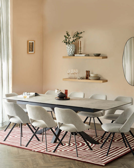 A modern dining room setting featuring a sleek, rectangular table with a dark surface and black metal legs, surrounded by six mid-century-inspired upholstered dining chairs with light grey fabric and angled black metal legs. Above the table, two natural wood floating shelves display decorative items. The dining set is anchored on a geometric red and white patterned area rug.