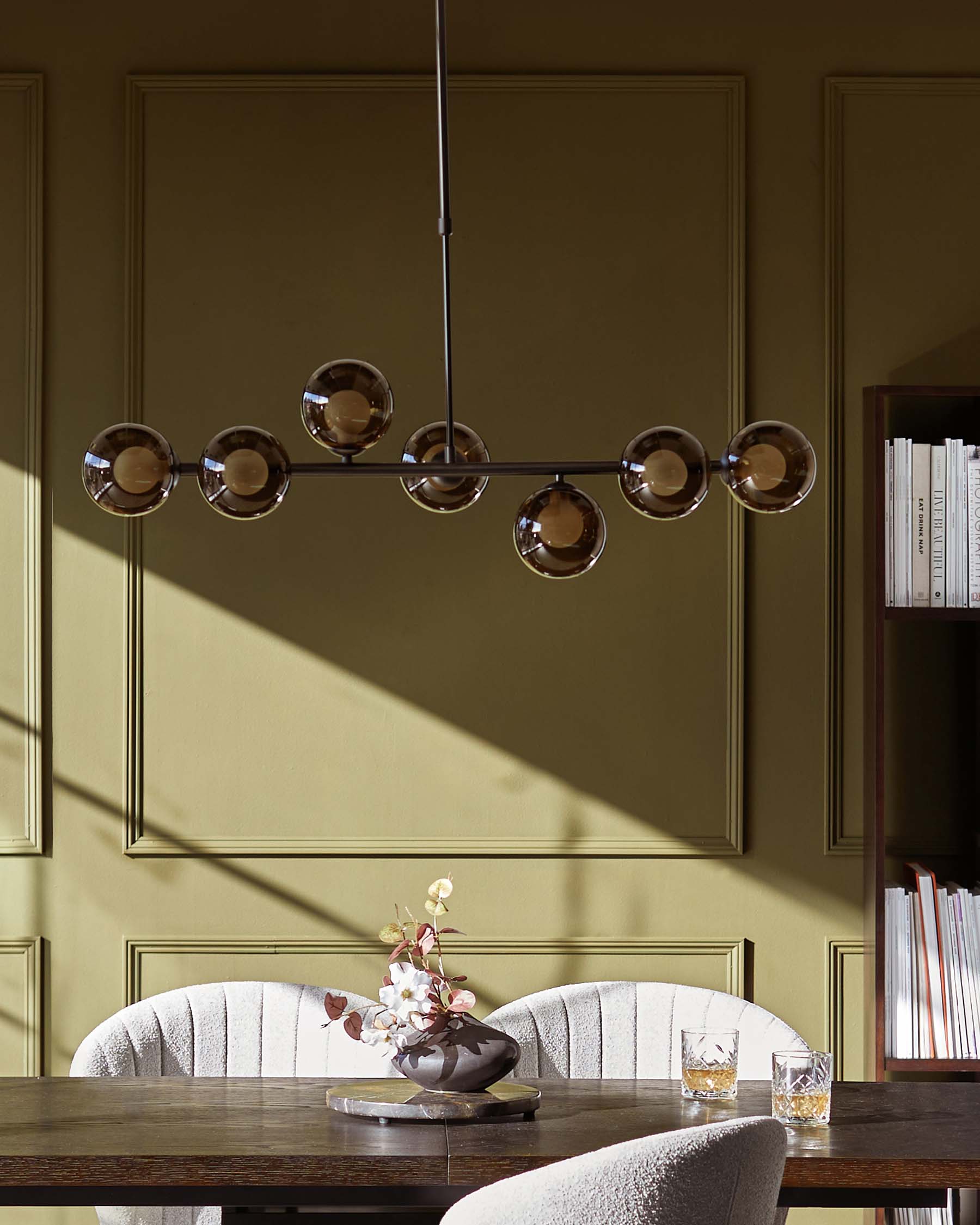 Elegant dining area featuring a modern rectangular wooden table with a rich, dark finish, complemented by two cosy, ribbed velvet upholstered chairs in a soft neutral tone with a subtly arched back design. A sophisticated, geometric chandelier with spherical glass shades hangs overhead, while natural light casts shadows on the panelled olive-green wall, enhancing the room's warm ambiance. A decorative floral arrangement and two glasses rest atop the table, completing the scene.