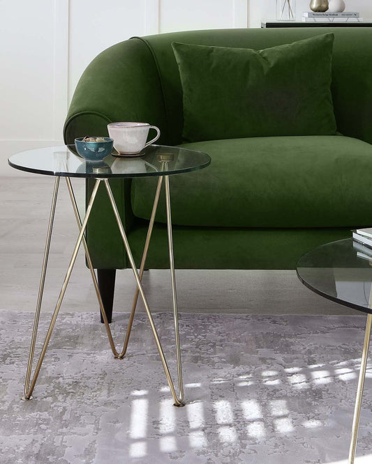 A green velvet sofa with a throw pillow, paired with elegant glass side tables featuring gold legs and decorative dishes.