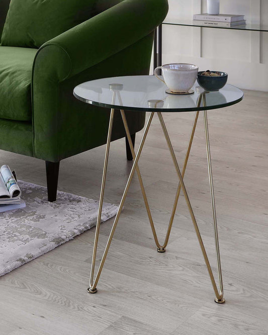 A modern glass-top side table with gold legs, accompanied by a cup and a small bowl, next to a green velvet sofa.