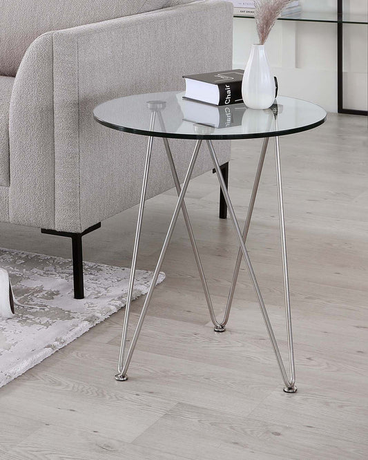 Round glass side table with sleek metal legs, accompanied by books and a decorative vase, displayed next to a modern sofa.