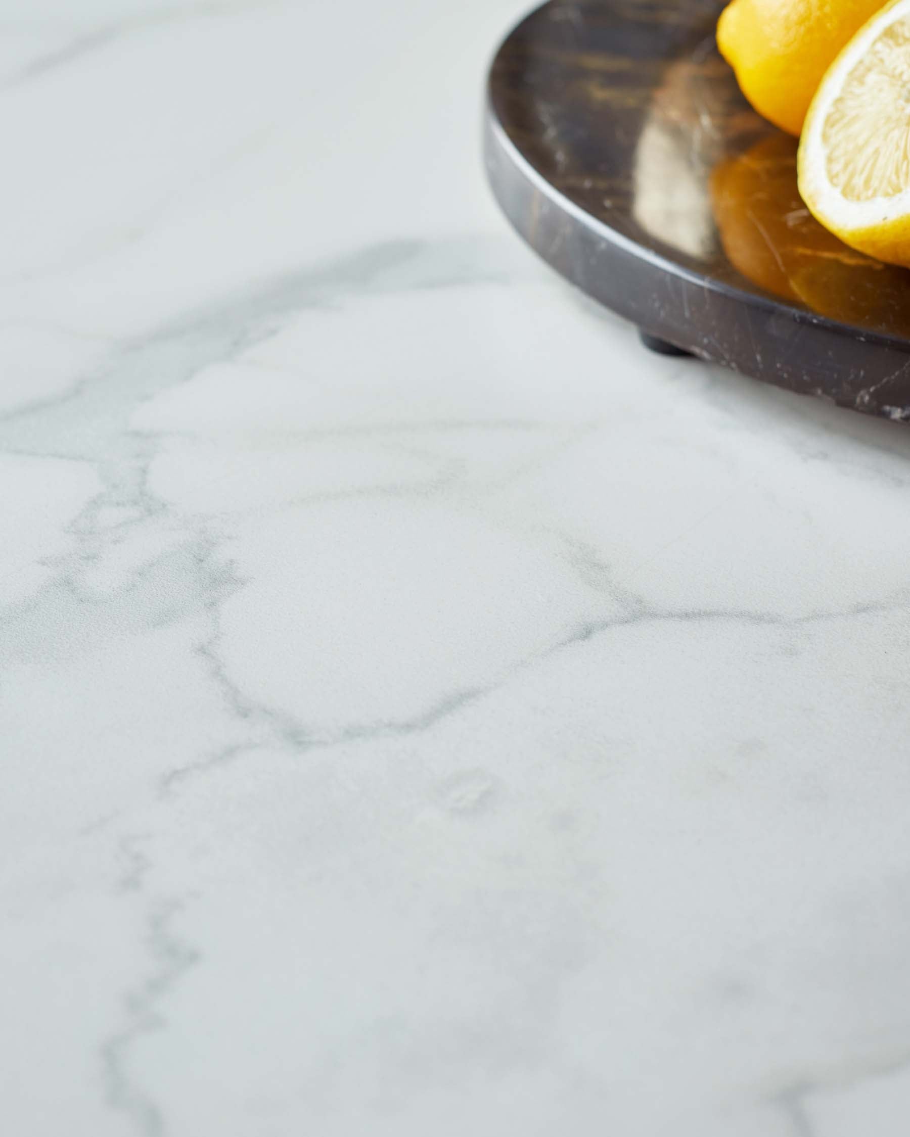 A smooth, light marble table surface with subtle veins, featuring a round dark platter holding fresh lemons.