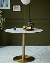 Round marble-top dining table with a gold metal pedestal, adorned with a French press, fruit, and a textured rug underneath.