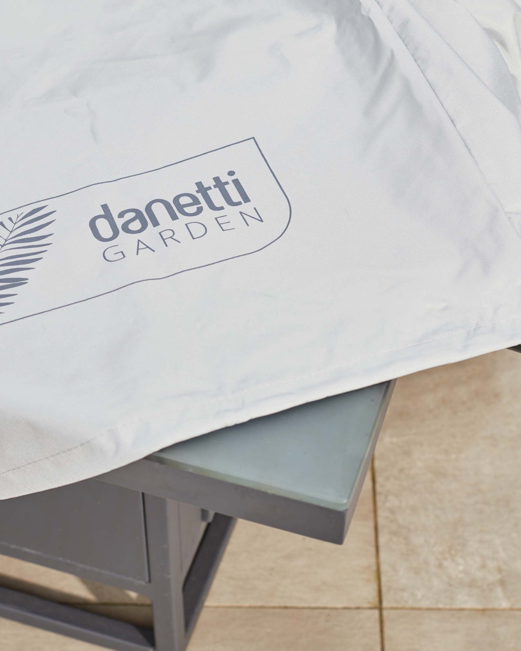 Close-up of a modern outdoor furniture cushion with "Danetti Garden" logo on white fabric, partially covering a glass-topped metal garden table.