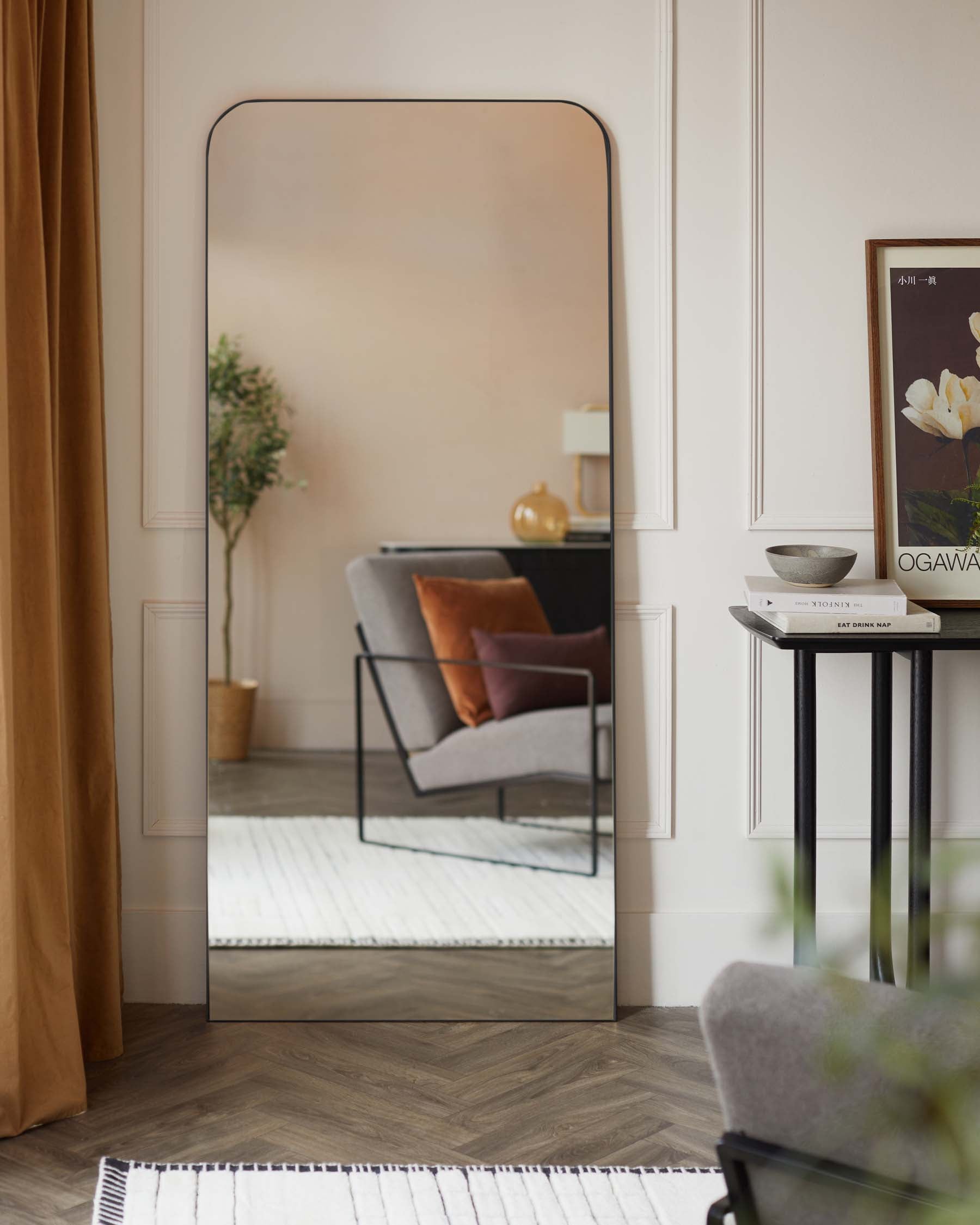 A large, free-standing floor mirror with a rounded top and thin black frame set against a neutral wall. A round side table with a black metal frame and marble top holds decorative items including a bowl, books, and a small vase. A glimpse of a grey armchair with a darker grey cushion and burnt orange pillow is reflected in the mirror.