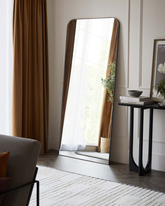 A tall, freestanding arched mirror with a wooden frame rests against a neutral wall, next to a slim, modern console table with a black metal frame and a dark tabletop. The table is styled with minimalist decor including a small bowl, books, and a framed artwork.