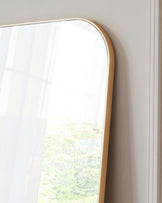 Modern rectangular mirror with rounded corners and a thin gold-coloured frame, leaning against a white wall with a partial reflection of a bright window and foliage outside.