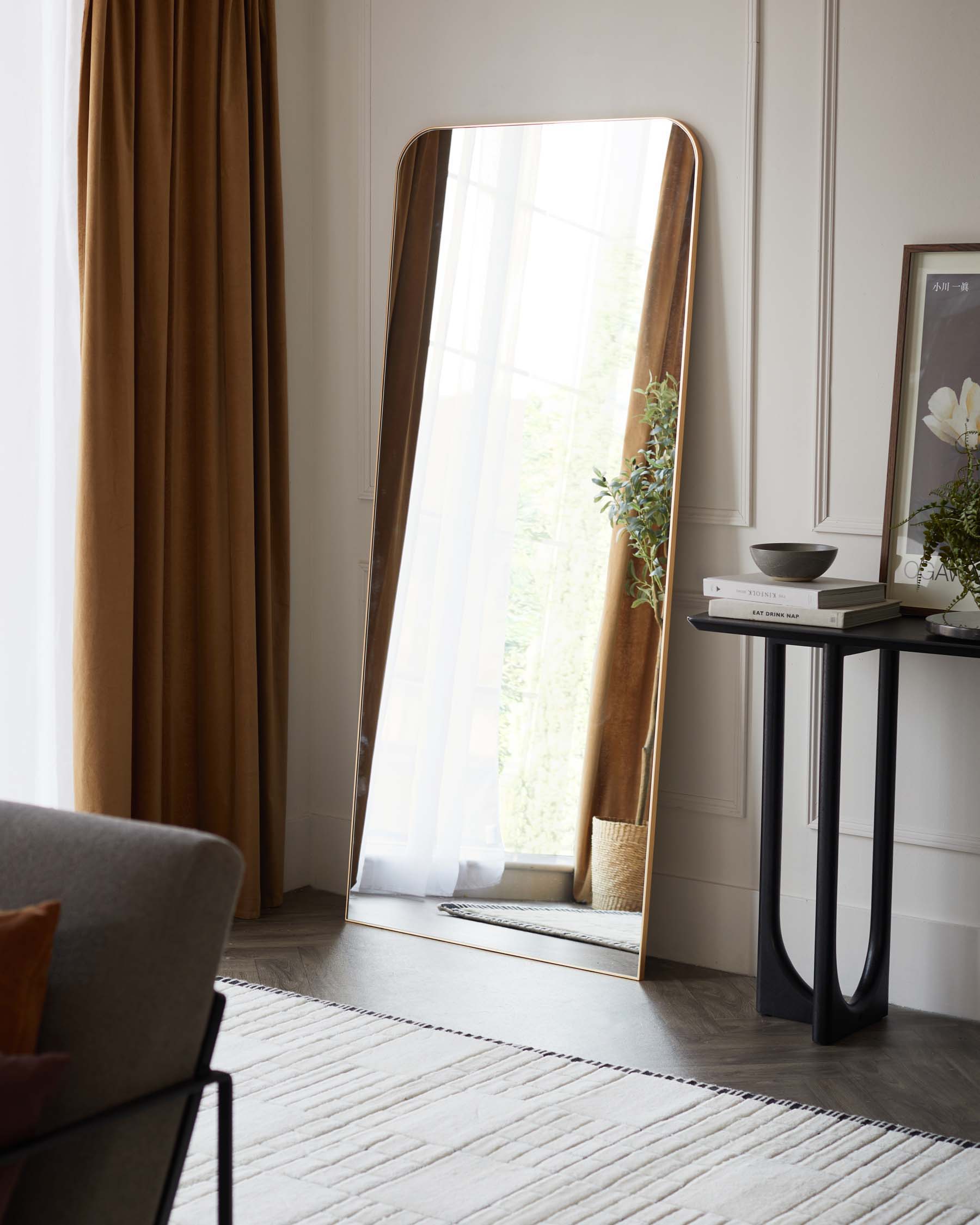 An elegant, full-length floor mirror with a gently curved top and a slim metallic frame stands against a neutral wall, casting reflections of the room's natural light. To the mirror's right, a minimalist black console table with slender, curved legs and a smooth surface displays decorative items, demonstrating a blend of functionality and modern aesthetics.