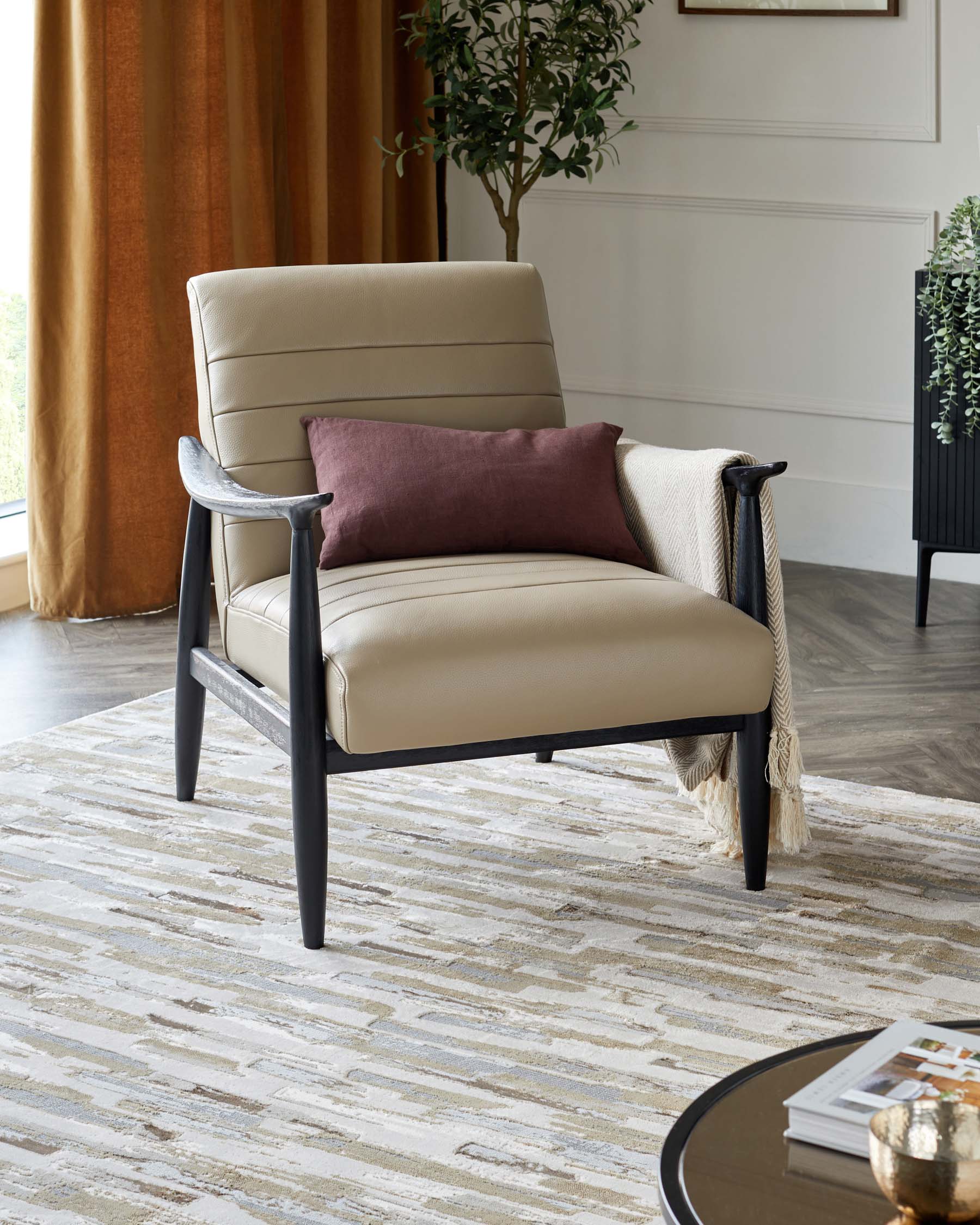 Modern beige leather armchair with a high backrest and sleek armrests set in a matte black frame. The chair is accessorized with a burgundy throw pillow and a cream tasselled blanket. The room features a textural beige and grey rug, a round black coffee table with a glossy finish, and a potted green plant beside a black shelving unit. Warm golden curtains and white panelled walls create an elegant background.