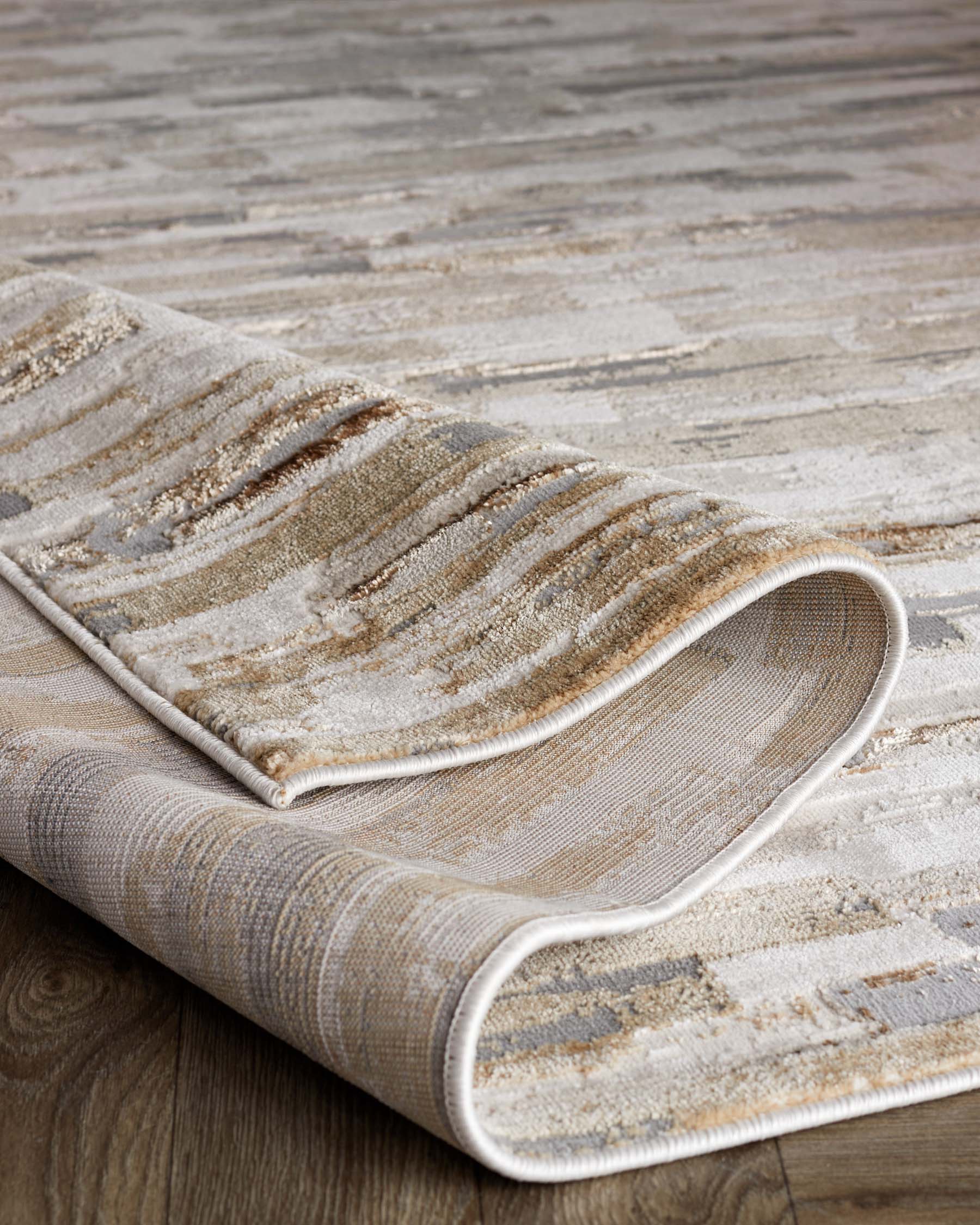 Elegant textured area rug with a rolled edge, featuring a blend of neutral tones with hints of beige and grey, laid on a wooden floor.