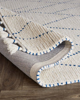 Handwoven area rug with beige base and blue accent lines, featuring intricate textures and fringe detailing, partially rolled to show underside on wooden flooring.