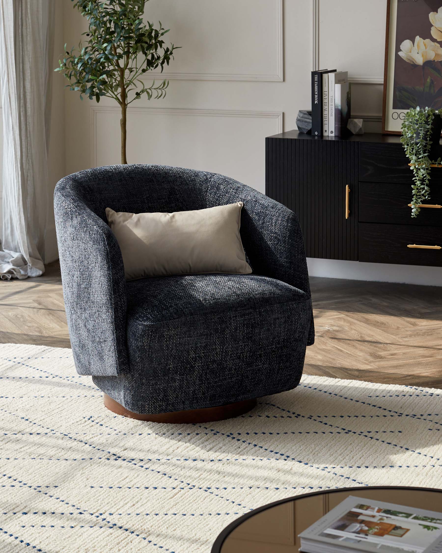 A contemporary dark grey upholstered armchair with a rounded back and a single beige cushion placed on a textured cream and blue-striped area rug. In the background, a modern black wood sideboard with vertical ridges and brass handles is partially visible, adorned with books, framed artwork, and greenery.