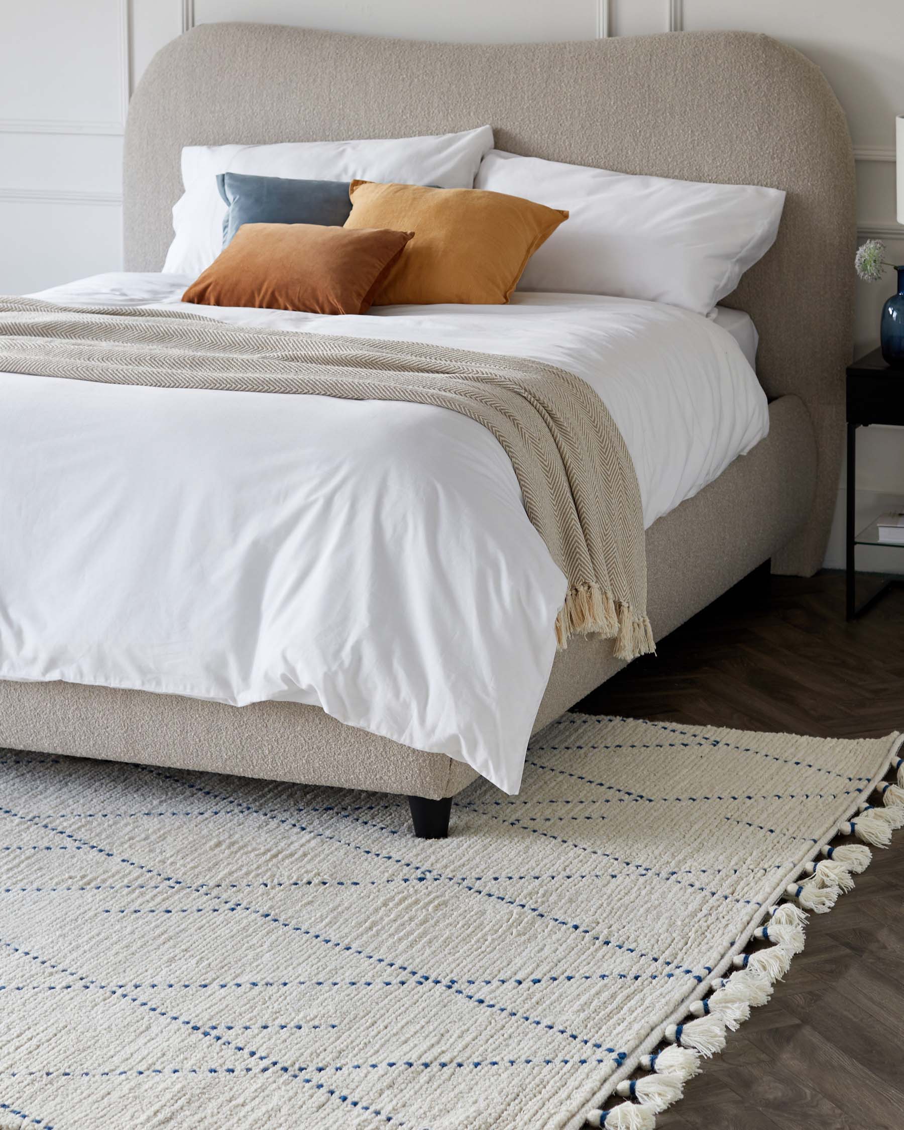 Contemporary upholstered platform bed with a curved headboard in a textured beige fabric, featuring a clean, white bedding set accentuated by mustard and slate blue pillows. At the foot of the bed is a white knit throw blanket. Positioned on a white and blue patterned area rug with tassel detailing, the design is complemented by a black minimalist bedside table with a dark vase and foliage to the side.