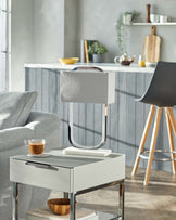 Modern minimalist furniture in a contemporary living room setting, featuring a light grey upholstered armchair with a smooth fabric finish, a sleek white side table with a glossy surface and metallic accents, and a tripod-style wooden floor lamp with a unique silhouette and beige lampshade, all complemented by a cosy yet chic ambiance.