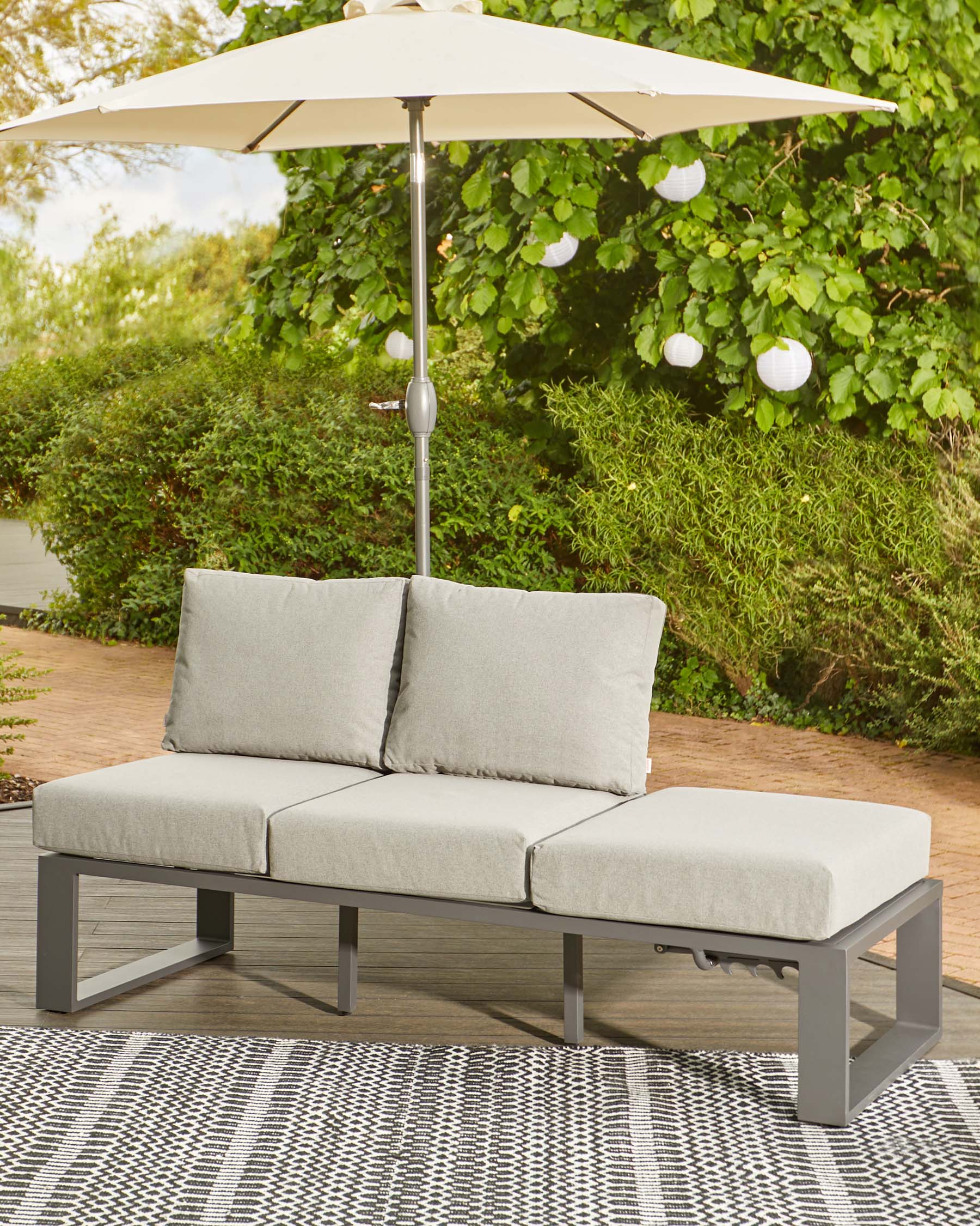 Gray outdoor sofa with plush cushions and an umbrella, set on a patterned rug in a lush garden backdrop.