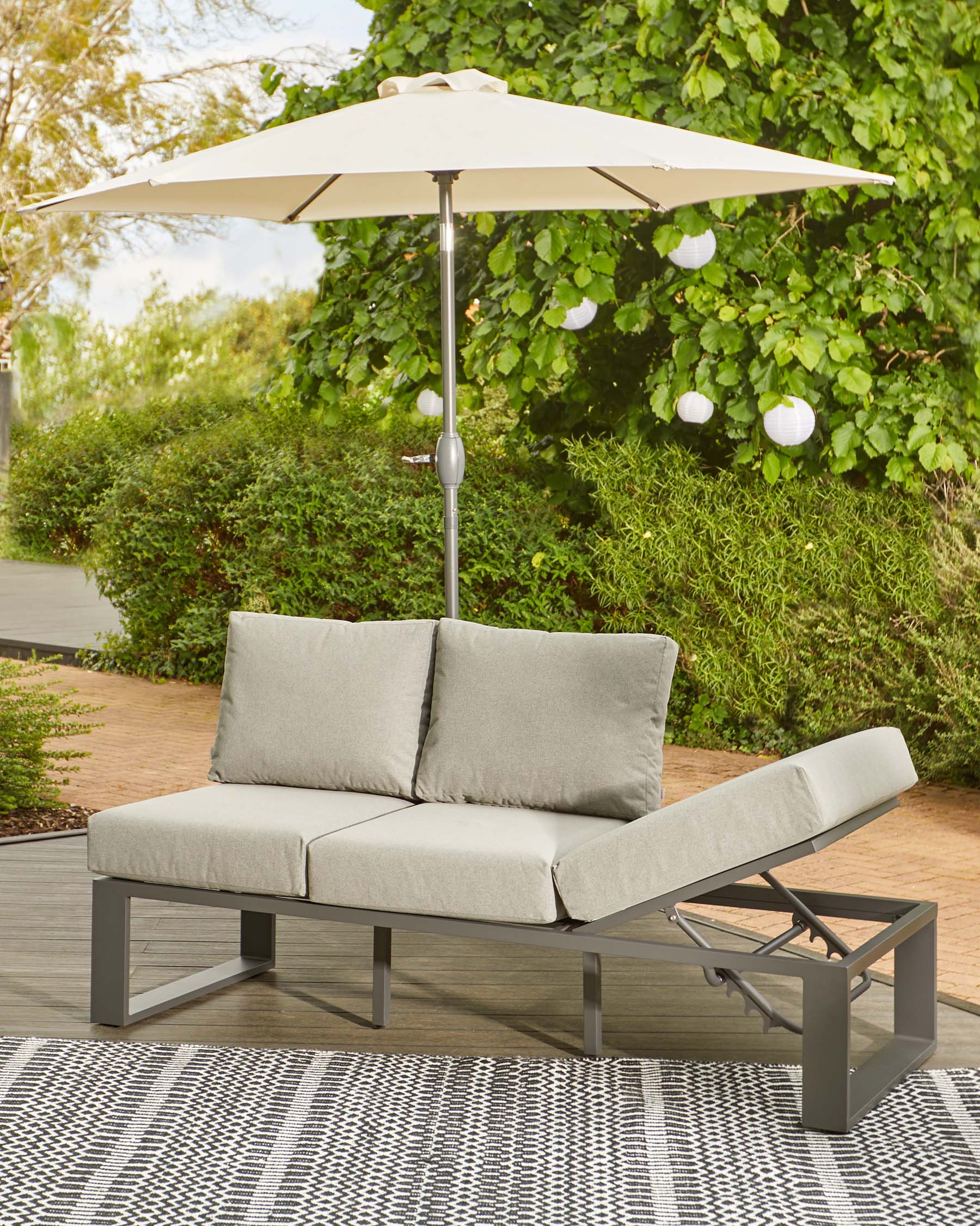 Modern outdoor sofa with gray cushions, adjustable backrest, and a large umbrella for shade, set on a patterned rug.