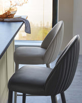 Two stylish upholstered bar stools with curved backs, one in light gray and the other in dark gray, set against a bright view.