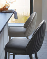 Two stylish upholstered bar stools with ribbed backs, one light gray and one darker gray, beside a modern kitchen countertop.