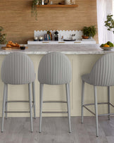 Three modern gray upholstered bar stools with ribbed design and sleek metal legs, set against a stylish kitchen backdrop.