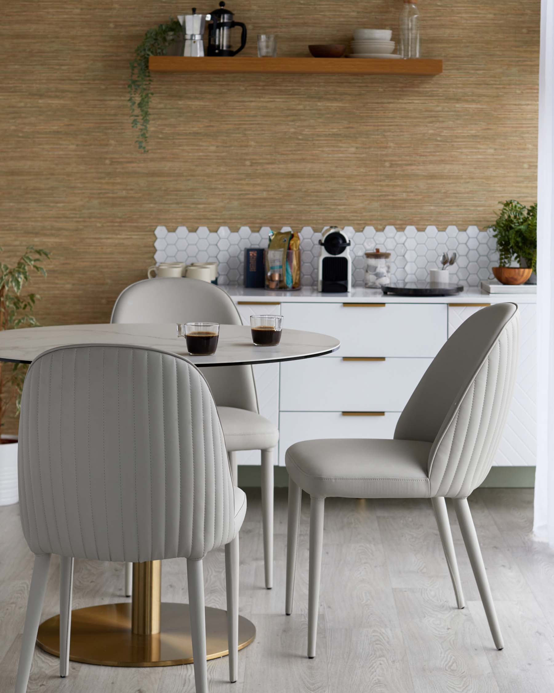 A round glass table with a gold base and three gray upholstered chairs featuring ribbed designs, set in a stylish kitchen.